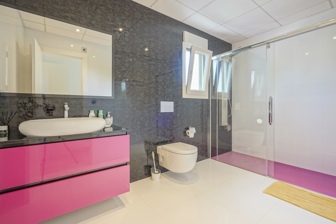 Moderne badkamer met een zwevend roze wastafelmeubel en een witte opzetwastafel. Voorzien van een glazen inloopdouche met roze vloer en donkere wandtegels met een subtiel patroon.