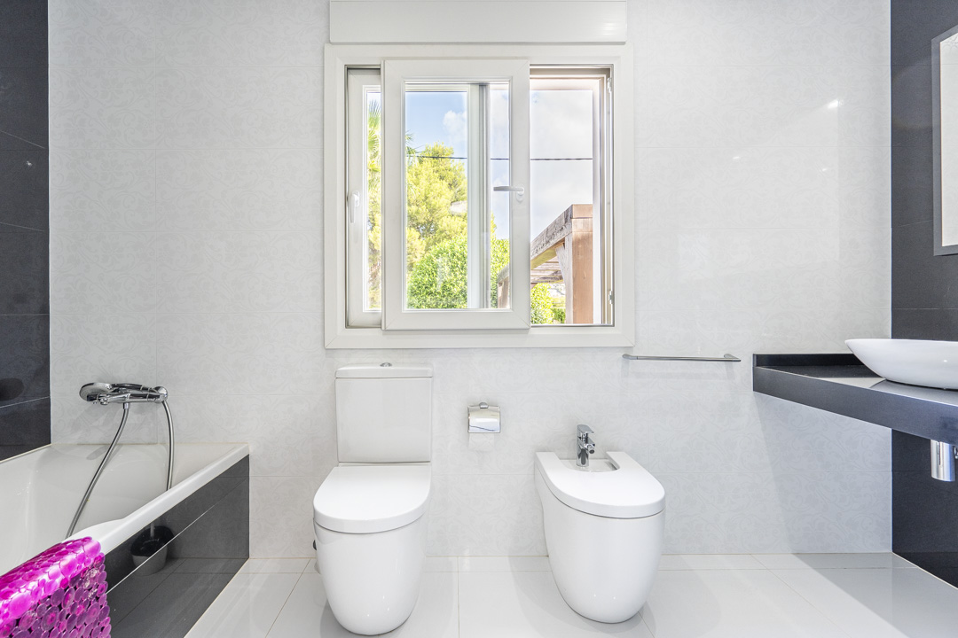 Moderne badkamer met toilet, bidet en ligbad. Voorzien van een zwart zwevend wastafelmeubel met opzetkom, witte reliëftegels en een raam met zicht op de tuin.