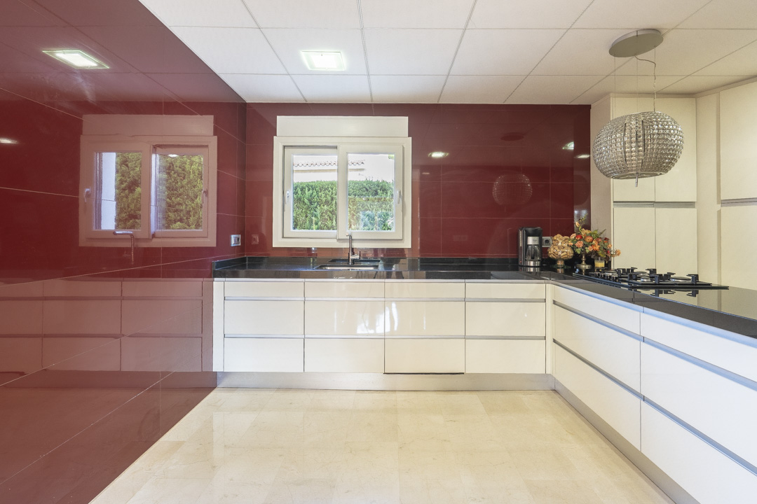 Moderne keuken met greeploze witte kasten, een zwart granieten werkblad en een opvallende rode achterwand. Voorzien van inbouwverlichting en een kristallen hanglamp.