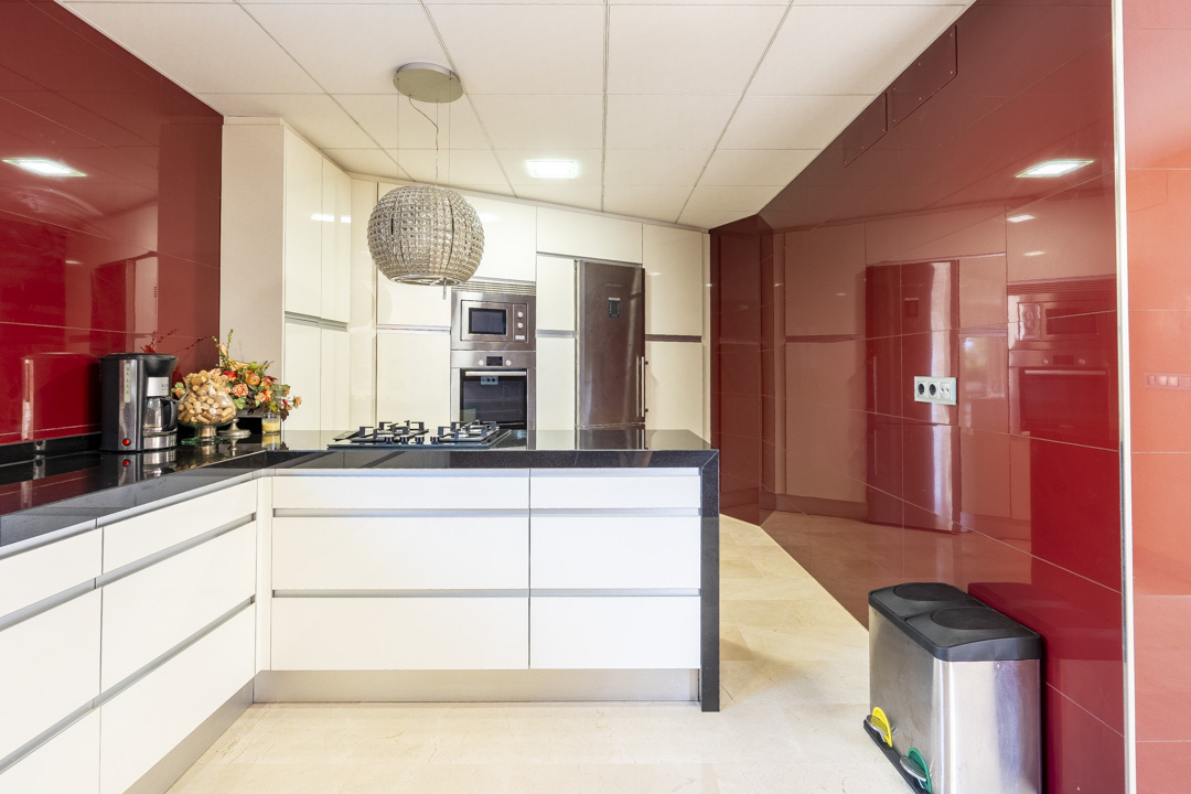 Moderne keuken met greeploze witte kasten, zwart granieten werkbladen en glanzend rode wandpanelen. Voorzien van inbouwapparatuur en een decoratieve bolvormige hanglamp.