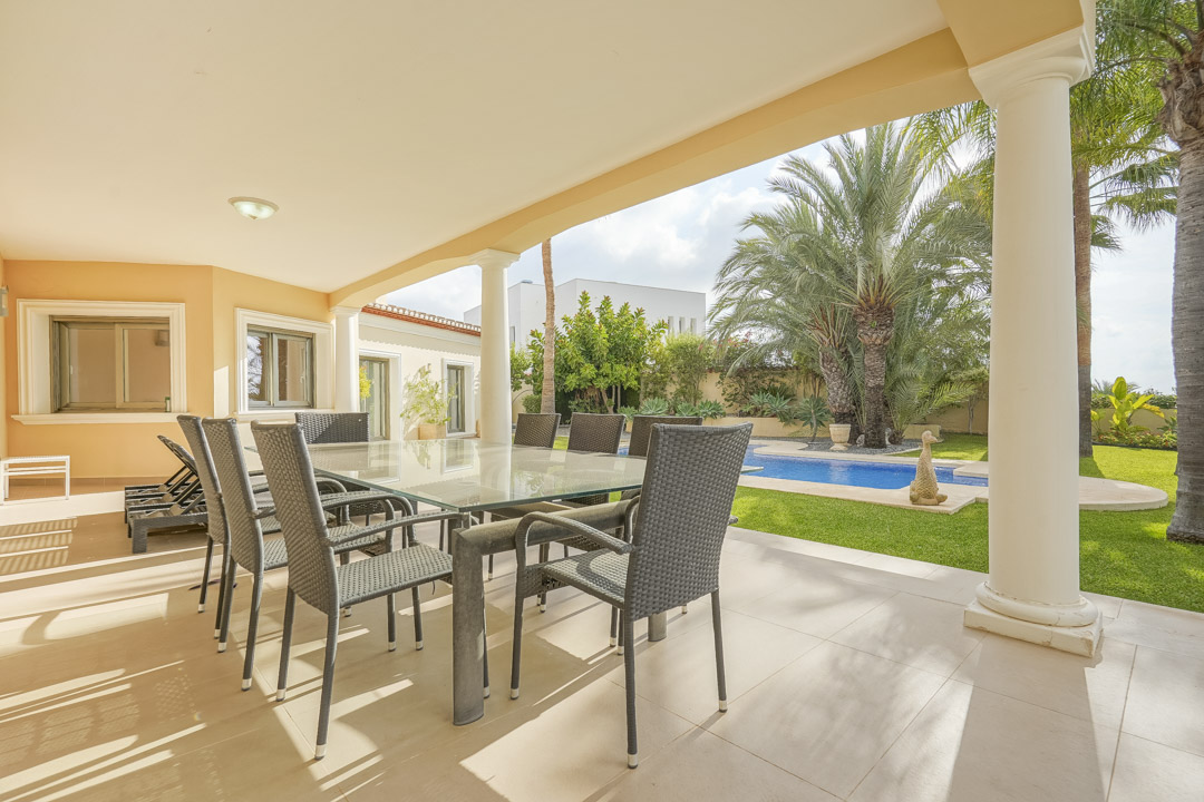 Overdekt terras met witte kolommen, glazen eettafel en uitzicht op het zwembad en de volgroeide mediterrane tuin.