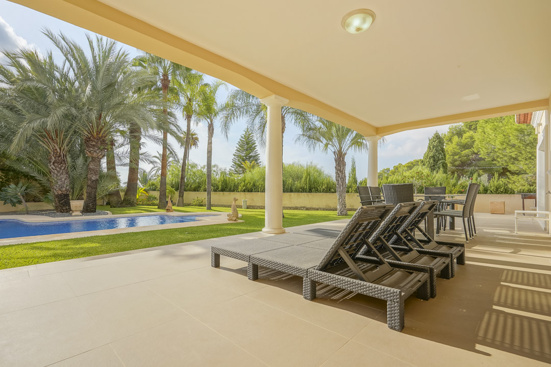 Ruim overdekt terras met witte zuilen en tuinmeubilair, uitkijkend op het zwembad en de volgroeide mediterrane tuin met diverse palmbomen.