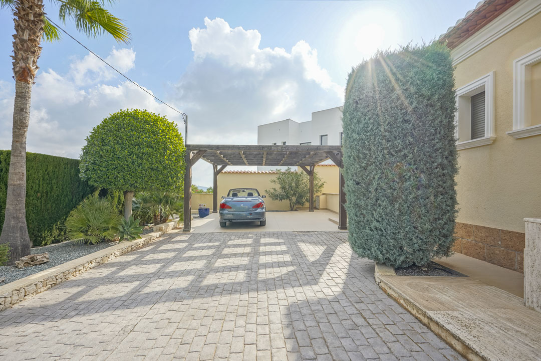 Ruime geplaveide oprit met een houten carport, omgeven door een volwassen mediterrane tuin en de lichte gevel van de villa.