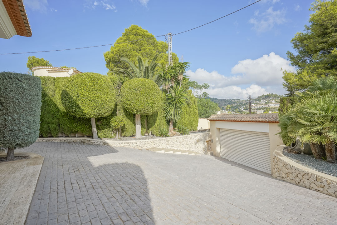 Ruime geplaveide oprit met toegang tot de garage, omgeven door een volgroeide mediterrane tuin met vormbomen en natuurstenen muren.