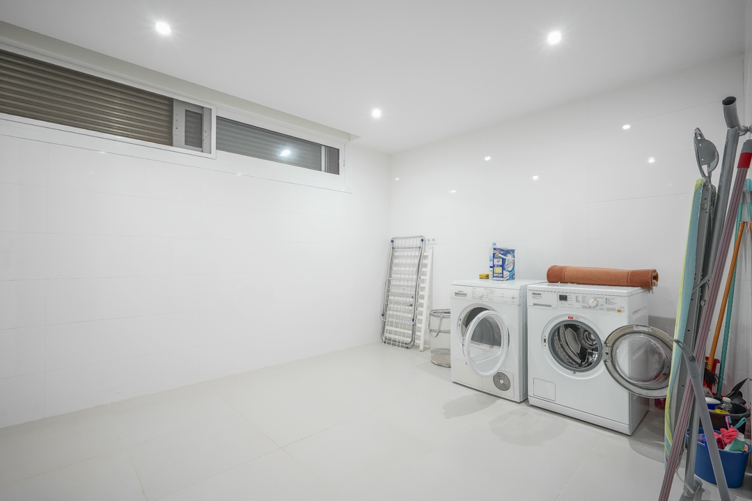 Ruime wasruimte in het souterrain met wit betegelde wanden, inbouwspots, een wasmachine en droger. Voorzien van een hoog geplaatst raam voor natuurlijke ventilatie.