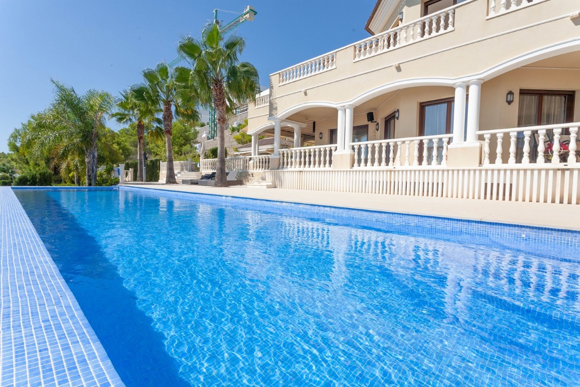 Blauw betegeld zwembad grenzend aan een mediterrane villa met gewelfde terrassen, witte balustrades en palmbomen onder een heldere hemel.