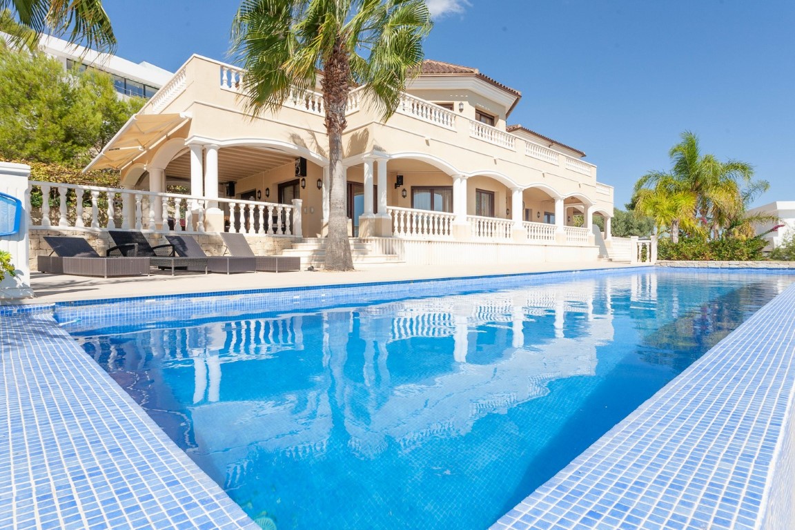 Groot zwembad met blauwe mozaïektegels bij een klassieke villa met overdekte boogterrassen, witte balustrades en mediterrane palmbomen.