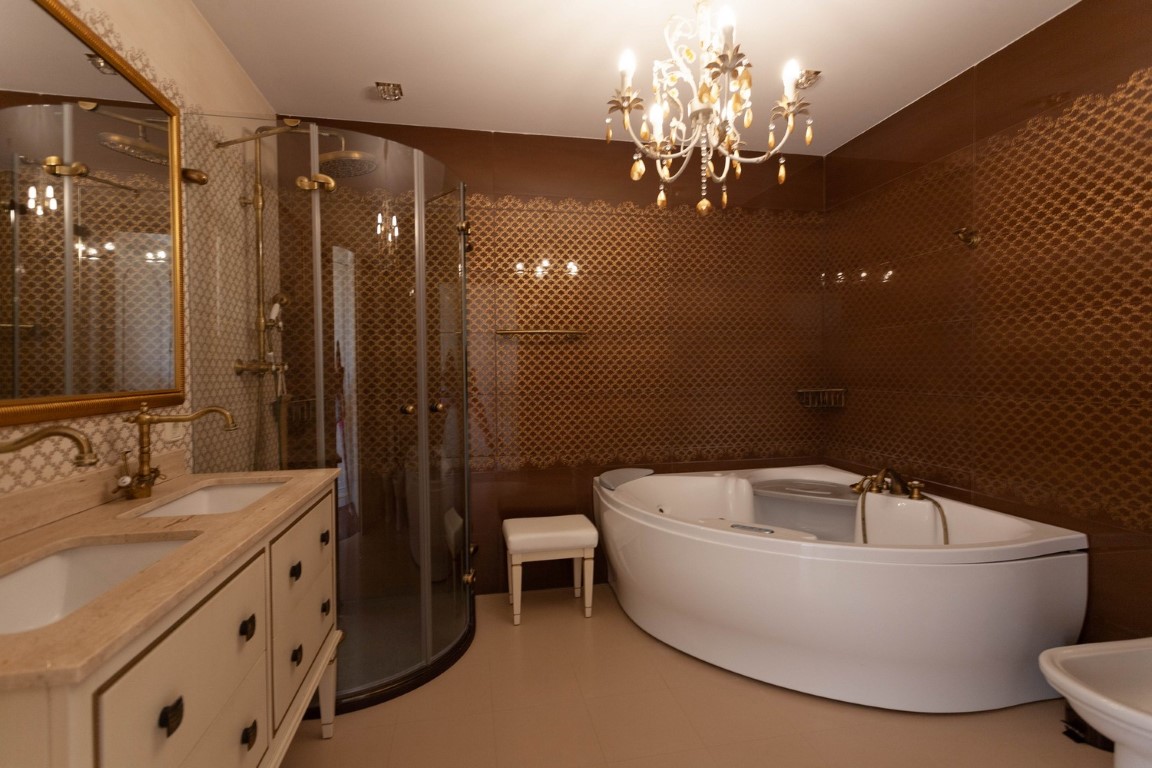Hoofdbadkamer met hoekwhirlpool, halfronde glazen douchecabine en dubbele wastafel met koperen kranen. De muren zijn afgewerkt met bruine structuurtegels en een klassieke kroonluchter.