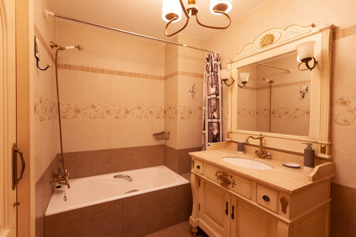 Klassieke badkamer met een sierlijk houten wastafelmeubel, stenen blad en bronzen kranen. Voorzien van een ingebouwd ligbad en decoratieve crème tegels.