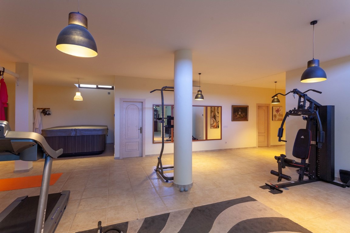 Privé fitness- en wellnessruimte met diverse trainingsapparatuur, een overdekte jacuzzi en tegelvloer, verlicht door industriële hanglampen.