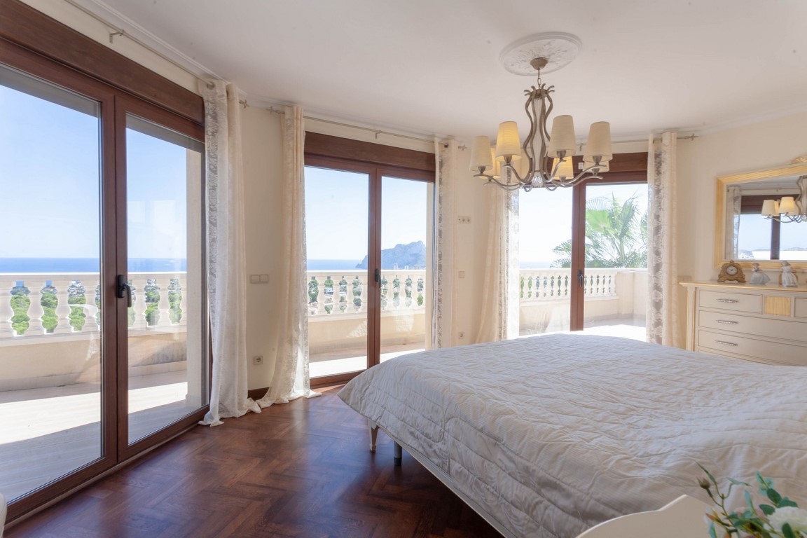 Slaapkamer met houten parketvloer, klassieke kroonluchter en grote schuifdeuren naar het balkon met uitzicht op de Middellandse Zee en de Peñón de Ifach.