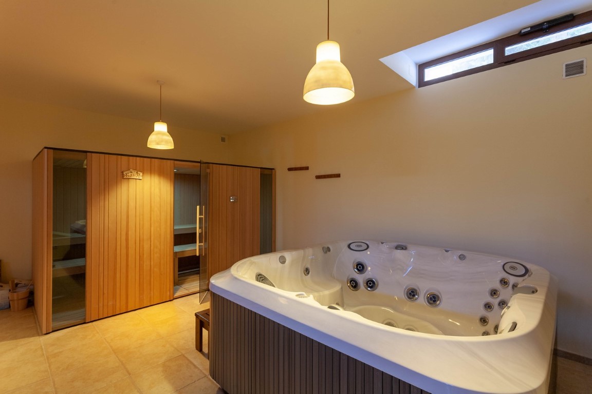 Wellnessruimte met een jacuzzi voorzien van massagejets en een houten sauna. De kamer heeft een tegelvloer, warme hanglampen en een hooggeplaatst raam voor lichtinval.