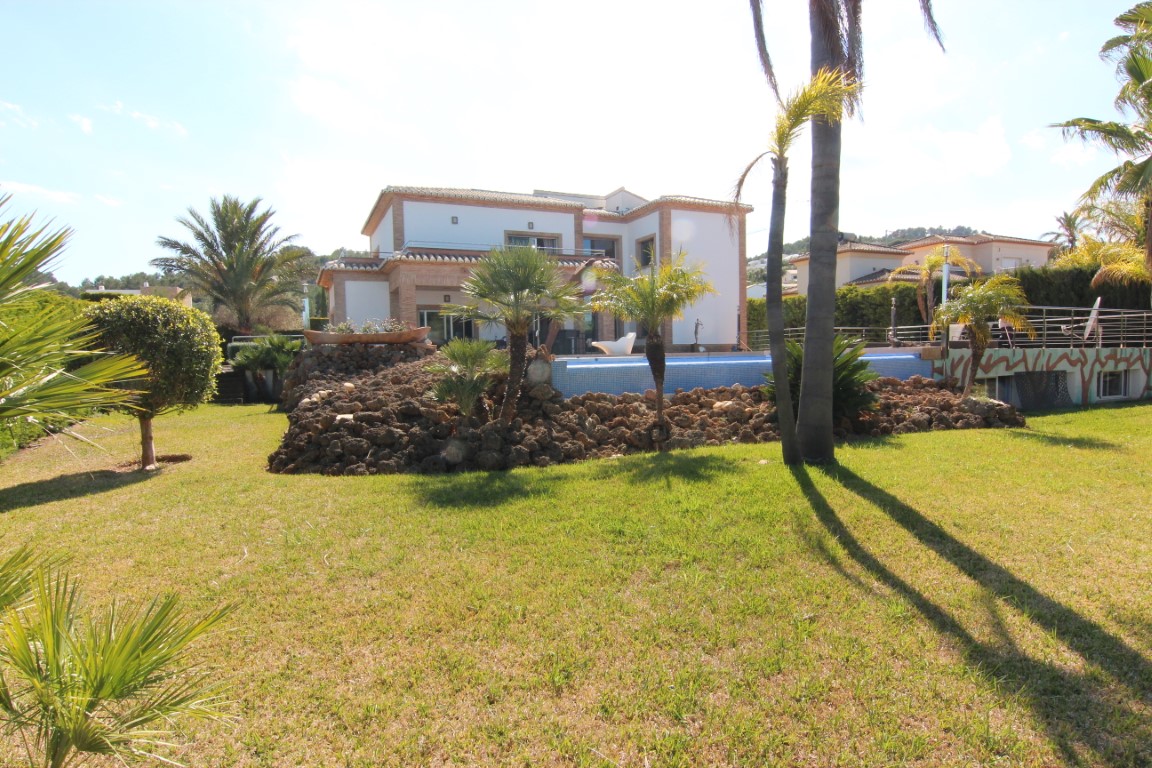 Buitenaanzicht van de villa met een verzorgd gazon, mediterrane beplanting en een verhoogd zwembad omgeven door natuurstenen rotspartijen en palmbomen.