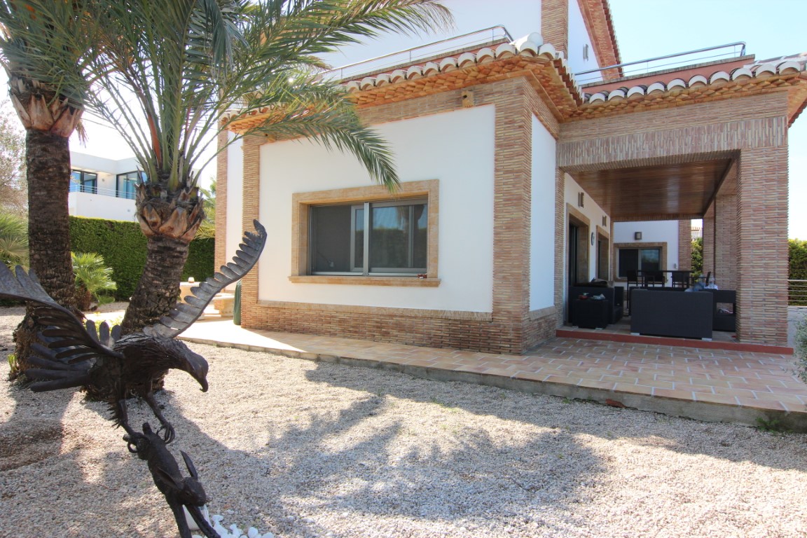 Exterieur van de villa met bakstenen details, een overdekt terras met tuinmeubilair en een grindtuin met palmbomen en een metalen adelaarsbeeld.