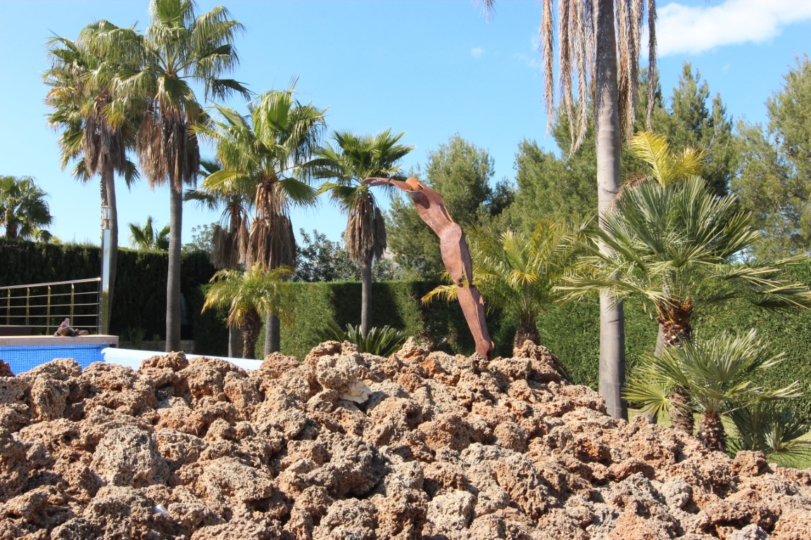 Mediterrane tuin met diverse palmbomen, een abstract metalen kunstwerk en natuurlijke rotsen bij het zwembad.