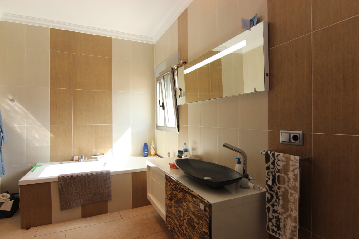 Moderne badkamer met ingebouwd ligbad, een design opzetwastafel en wandtegels in crème- en houtlook. Een grote spiegel met geïntegreerde verlichting verlicht de ruimte.