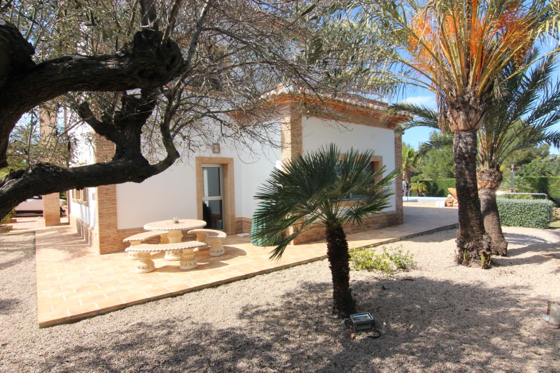 Zijterras met terracotta tegels en stenen meubilair, gelegen in een mediterrane tuin met palmbomen en volwassen bomen die natuurlijke schaduw bieden.