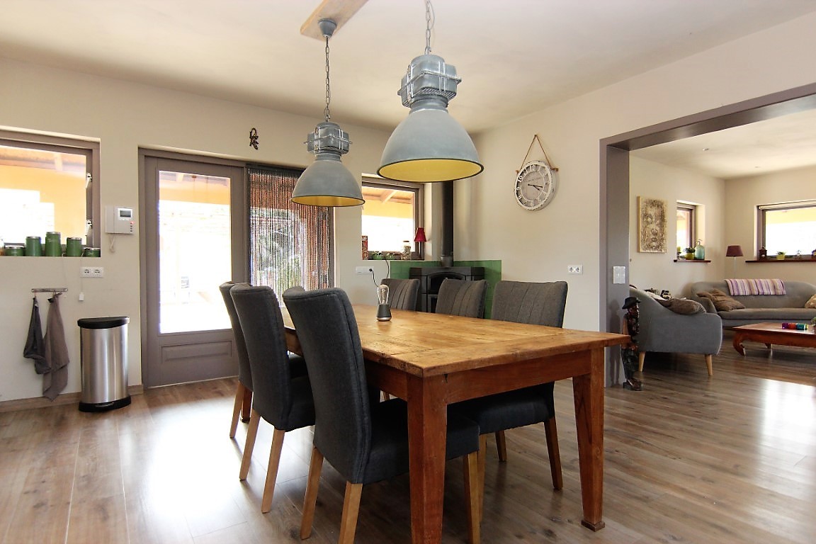 Eethoek met massief houten tafel, grijze gestoffeerde stoelen en industriële hanglampen, met een open verbinding naar de woonkamer en het terras.