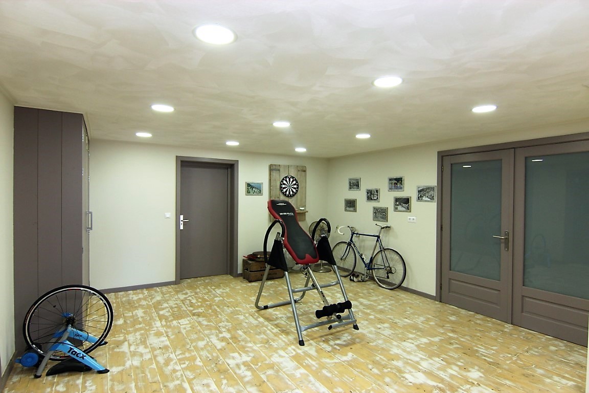 Fitnessruimte in het souterrain met lichte houten vloer, inbouwspots en ruimte voor sportuitrusting zoals fietsen en een inversiebank.