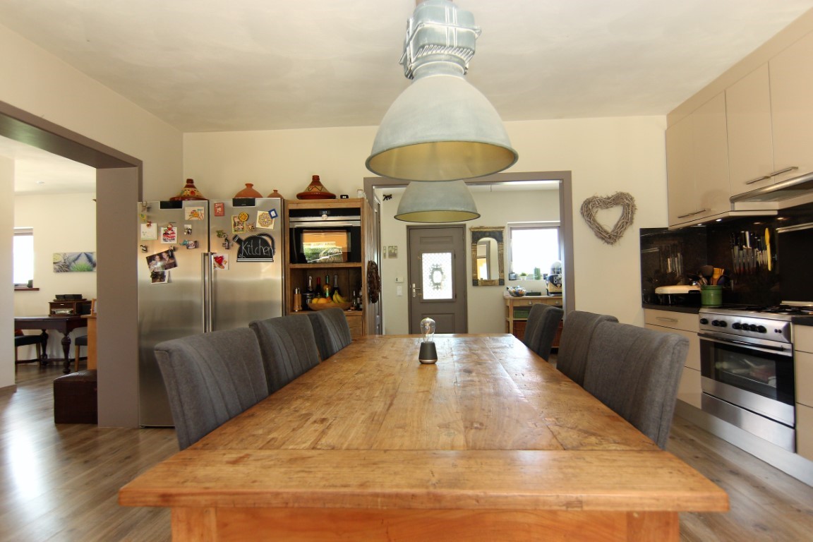 Keuken met eethoek voorzien van een grote rustieke houten tafel, industriële hanglampen en moderne roestvrijstalen inbouwapparatuur.