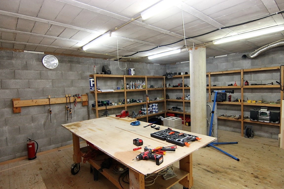 Ruime werkplaats in het souterrain met wanden van betonblokken, houten stellingkasten en een grote centrale werkbank op een houten vloer.