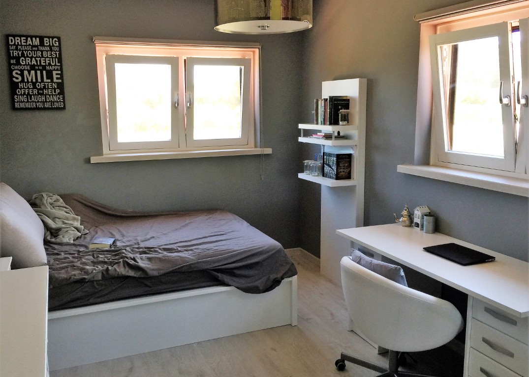 Tweepersoonskamer met een werkruimte voorzien van een wit bureau, grijze muren en een lichte houtvloer. Twee ramen bieden veel natuurlijk licht.