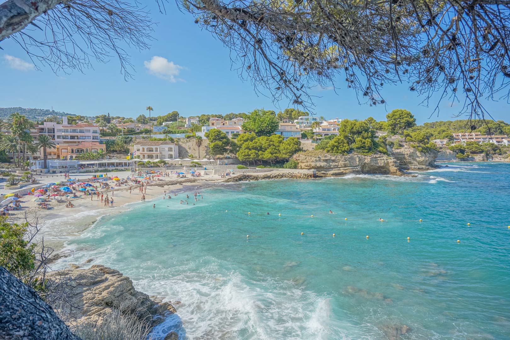 Panoramisch uitzicht op Cala de la Fustera met turquoise water, een zandstrand en kustwoningen, omlijst door mediterrane pijnbomen.