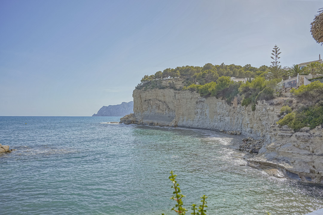 Panoramisch uitzicht op de kalkstenen kliffen en het turquoise water nabij het strand van La Fustera, met mediterrane pijnbomen en een rotsachtige kustlijn.