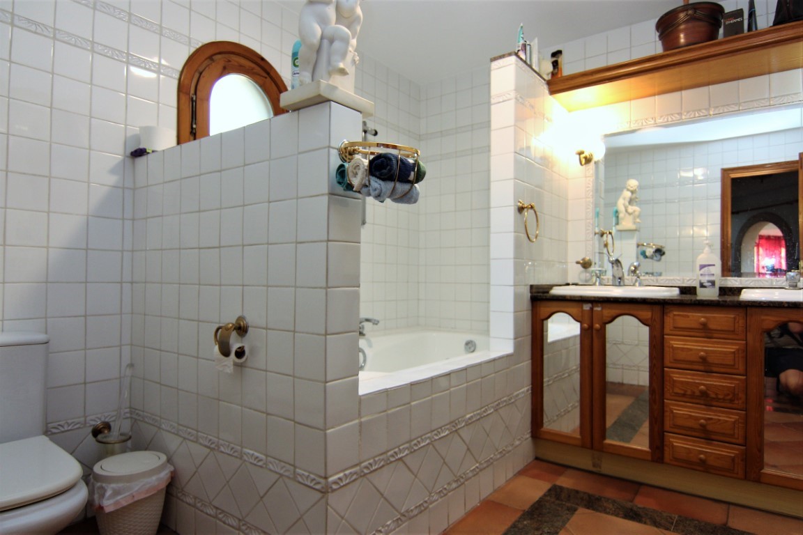 Badkamer met witte tegels, een ingebouwd ligbad en een houten dubbel wastafelmeubel. Voorzien van goudkleurige accenten, een boograam en een terracotta vloer.