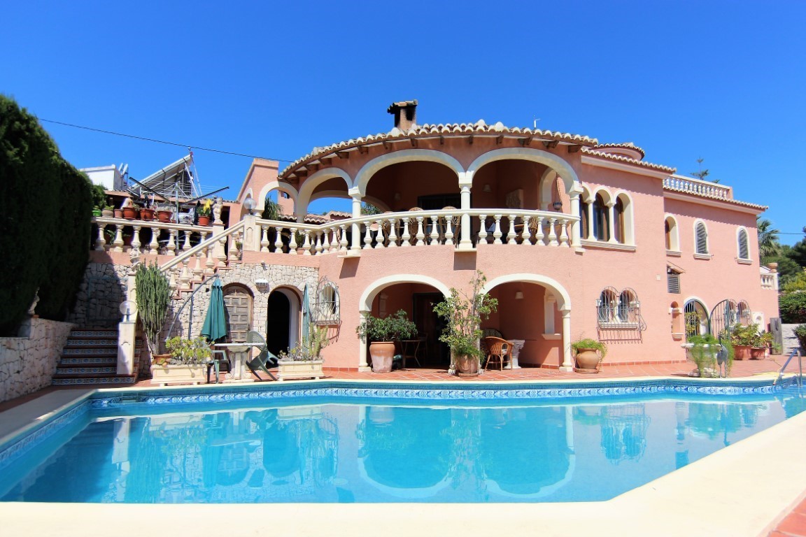 Buitenaanzicht van de villa met groot zwembad, zalmkleurige mediterrane gevel, overdekte terrassen met bogen en klassieke balustrades.