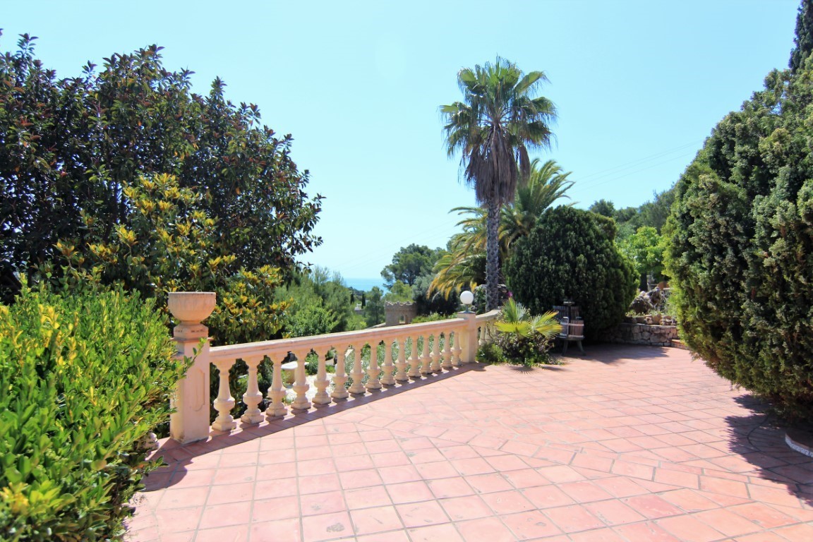 Buitenterras met terracotta tegels en een klassieke stenen balustrade, omgeven door volgroeide mediterrane beplanting met uitzicht op zee in de verte.