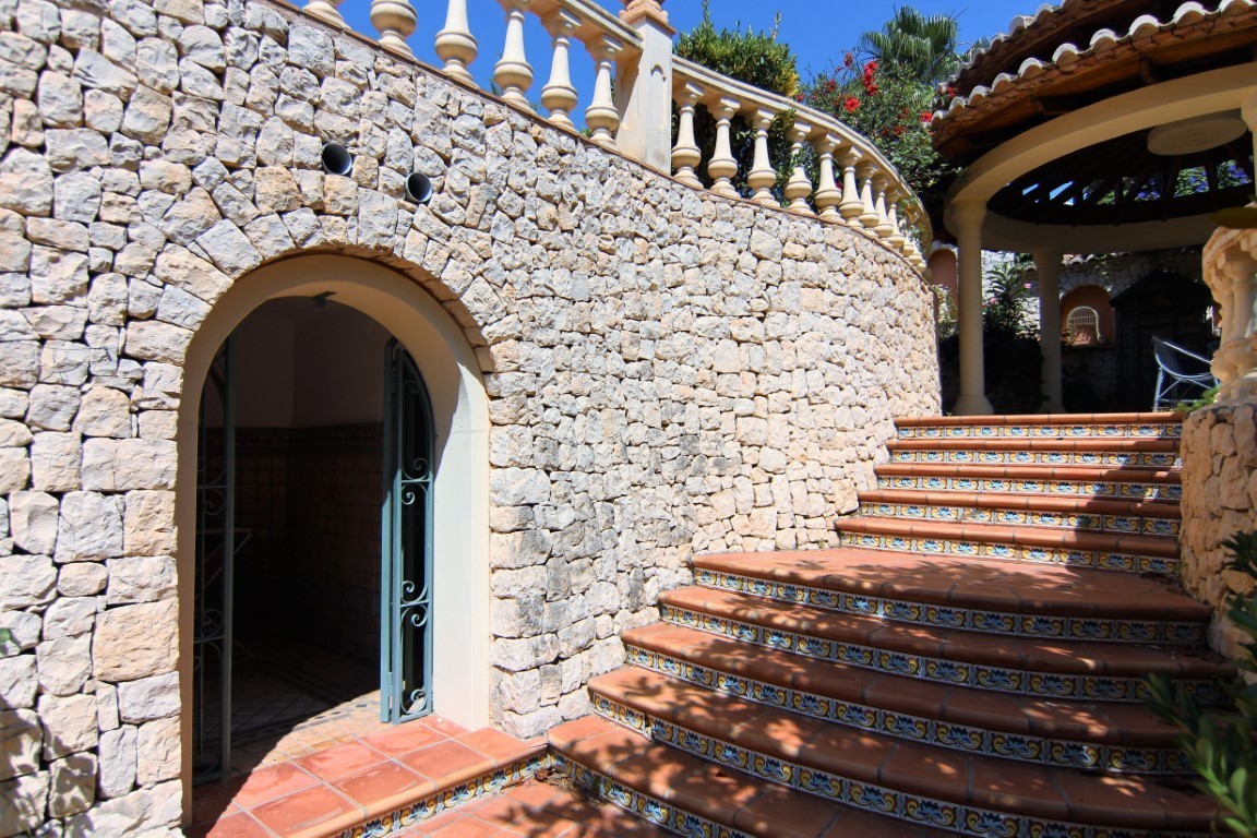 Buitentrap van terracotta met decoratieve tegels naast een gebogen natuurstenen muur. Voorzien van een rondboogingang en klassieke balustrade.