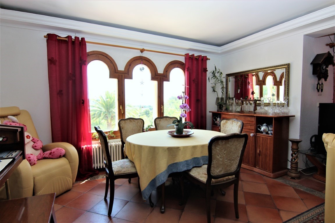 Eethoek met ronde tafel, gestoffeerde stoelen en een houten drieluik boogvenster. Voorzien van een terracotta tegelvloer en een houten dressoir met spiegel.