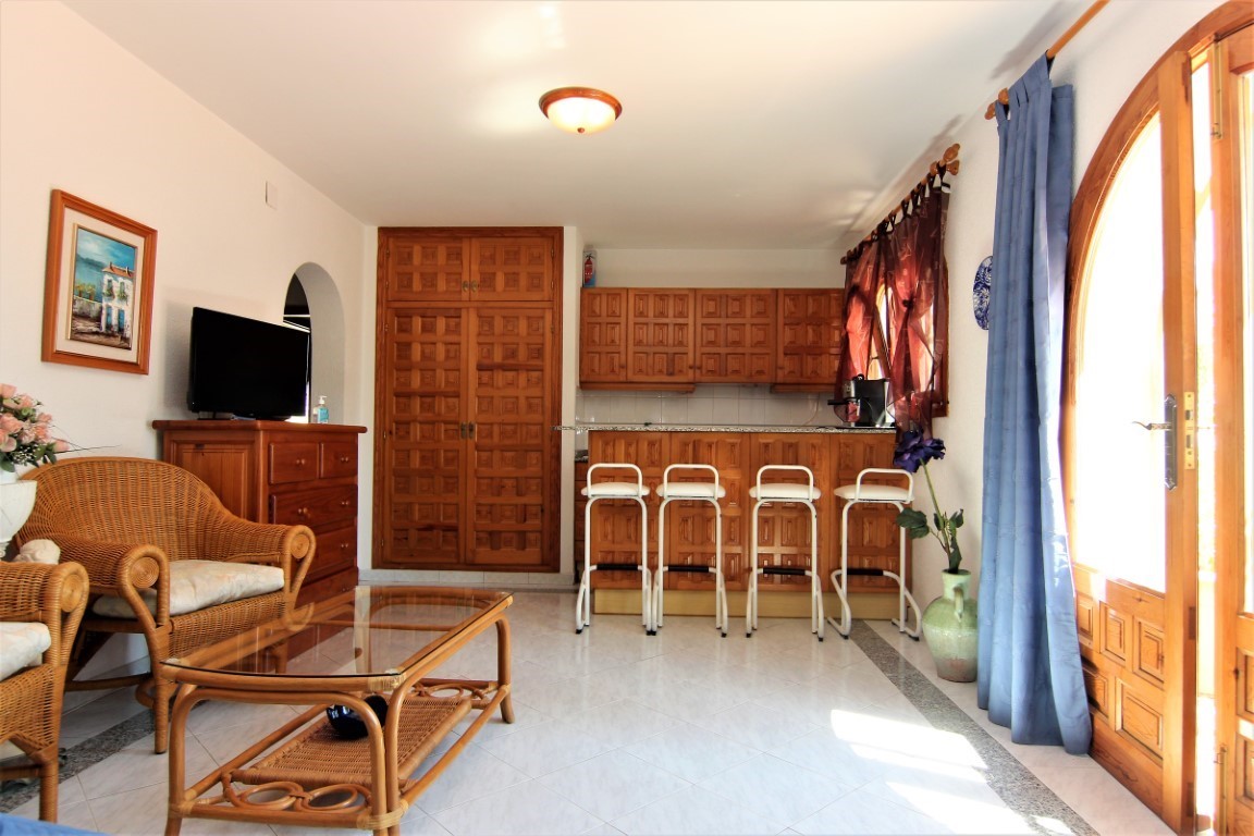 Gecombineerde woonkamer met kitchenette, rotan meubilair en houten inbouwkasten. De ruimte is voorzien van een tegelvloer en directe toegang tot buiten via boogdeuren.