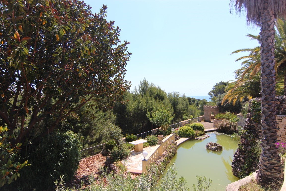 Grote siervijver met waterval, omgeven door volgroeide mediterrane beplanting en stenen paden met uitzicht op zee in de verte.