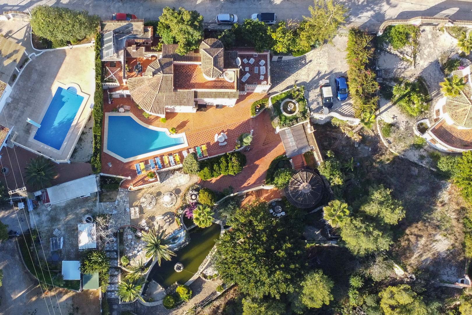 Luchtfoto van het perceel met het niervormige zwembad, uitgestrekte terracottaterrassen, een grote vijver met fontein en de volgroeide mediterrane tuin rondom de villa.