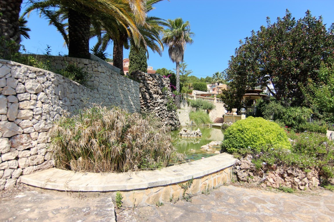 Mediterrane tuin met een siervijver, natuurstenen waterval en muren van gestapelde stenen, omgeven door palmbomen en weelderige beplanting.