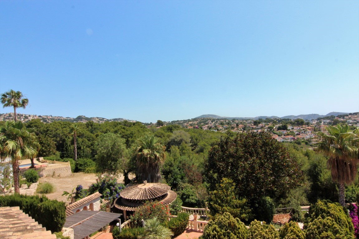 Panoramisch uitzicht vanaf het boventerras op de mediterrane tuin met palmbomen, een rond prieel met pannendak en de omliggende heuvels in de verte.