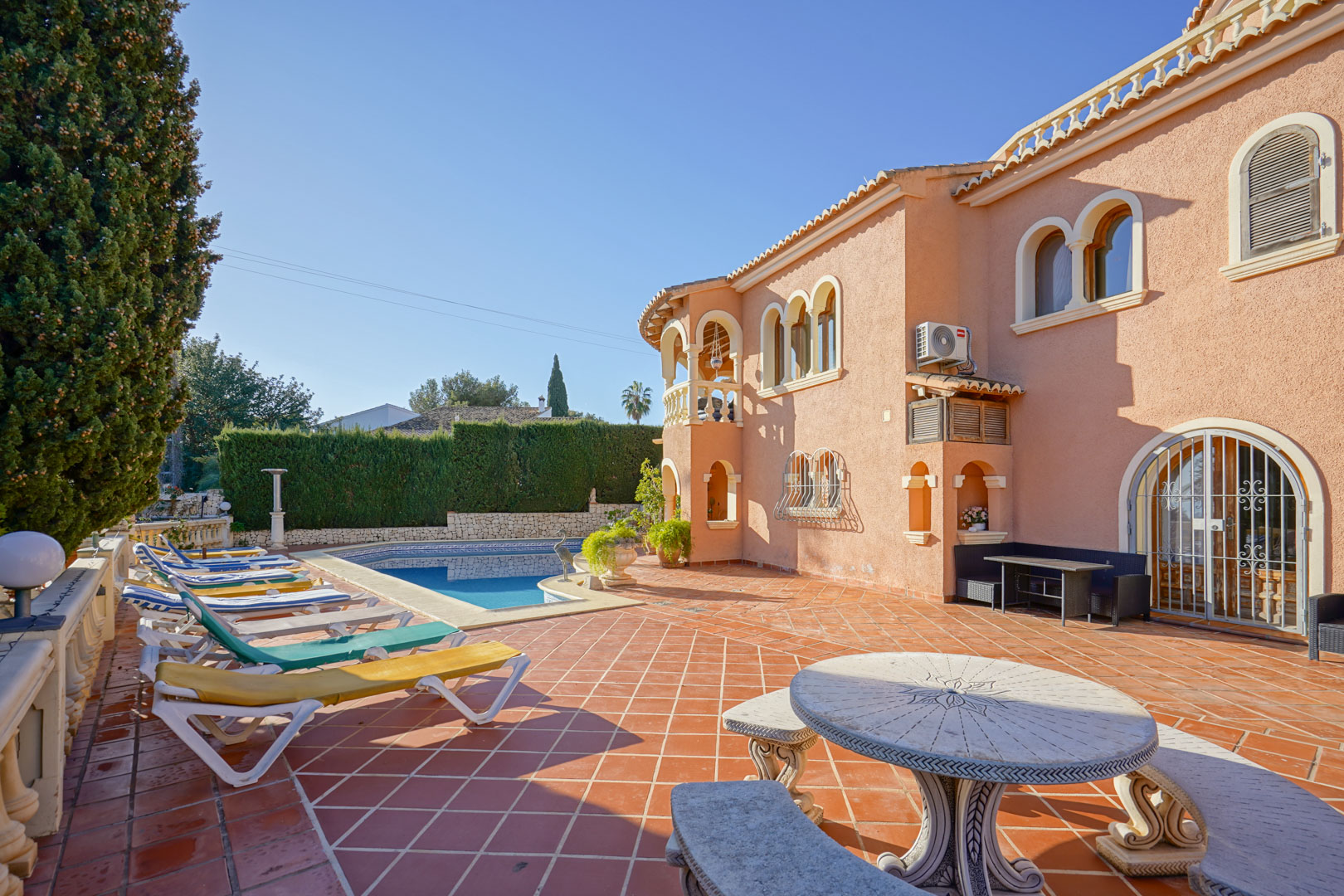 Ruim terras met terracotta tegels, een zwembad, diverse ligbedden en een bewerkte stenen tafel bij een mediterrane gevel met boogvensters.