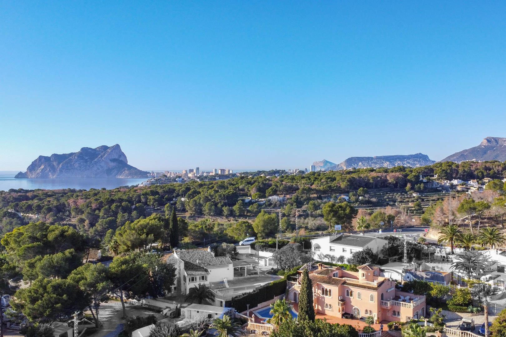 Verhoogd panoramisch uitzicht op de Middellandse Zeekust met de Peñón de Ifach, omgeven door dichte dennenbossen en residentiële villa's onder een heldere hemel.