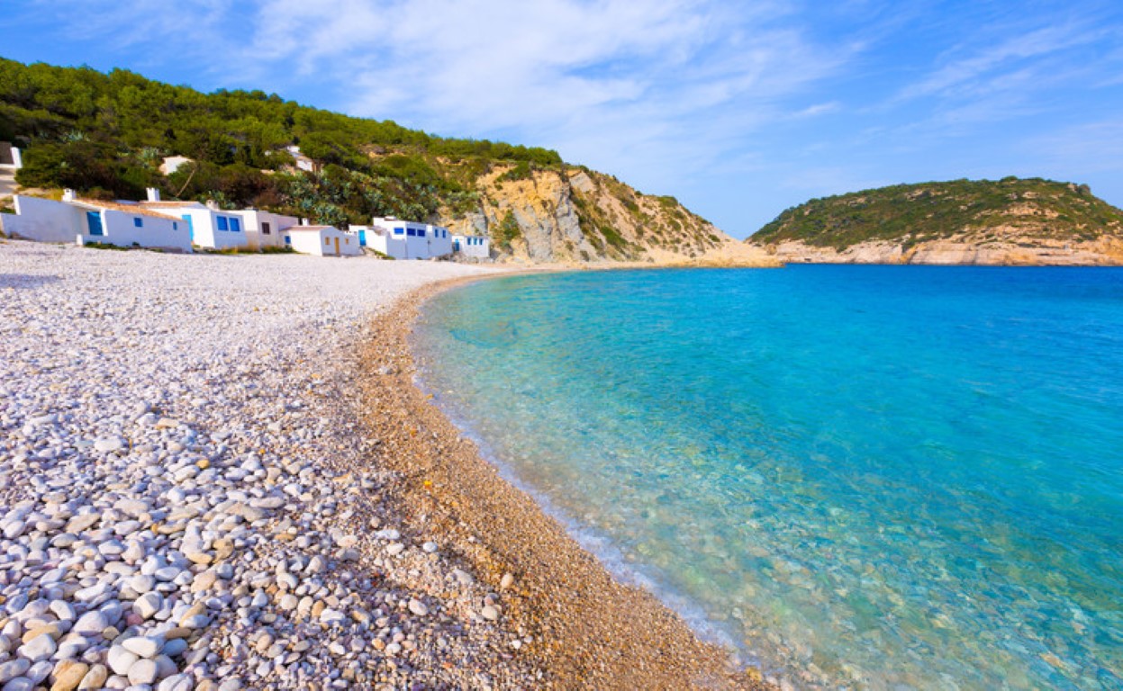 Uitzicht op Cala de la Barraca met turquoise water, wit kiezelstrand en karakteristieke vissershuisjes tegenover het eiland Portichol in Jávea.