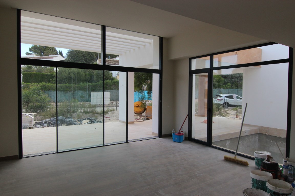 Woonkamer in afbouwfase met grote raampartijen in zwarte kozijnen die toegang geven tot het terras met een witte pergola-overkapping.
