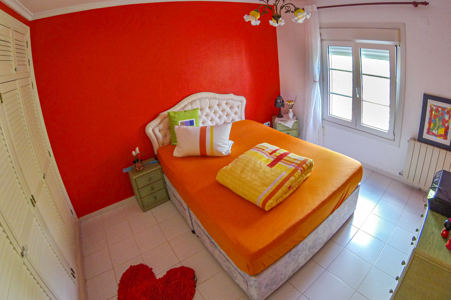 Lichte slaapkamer met rode accentmuur, tweepersoonsbed met oranje beddengoed en gestoffeerd hoofdeinde.