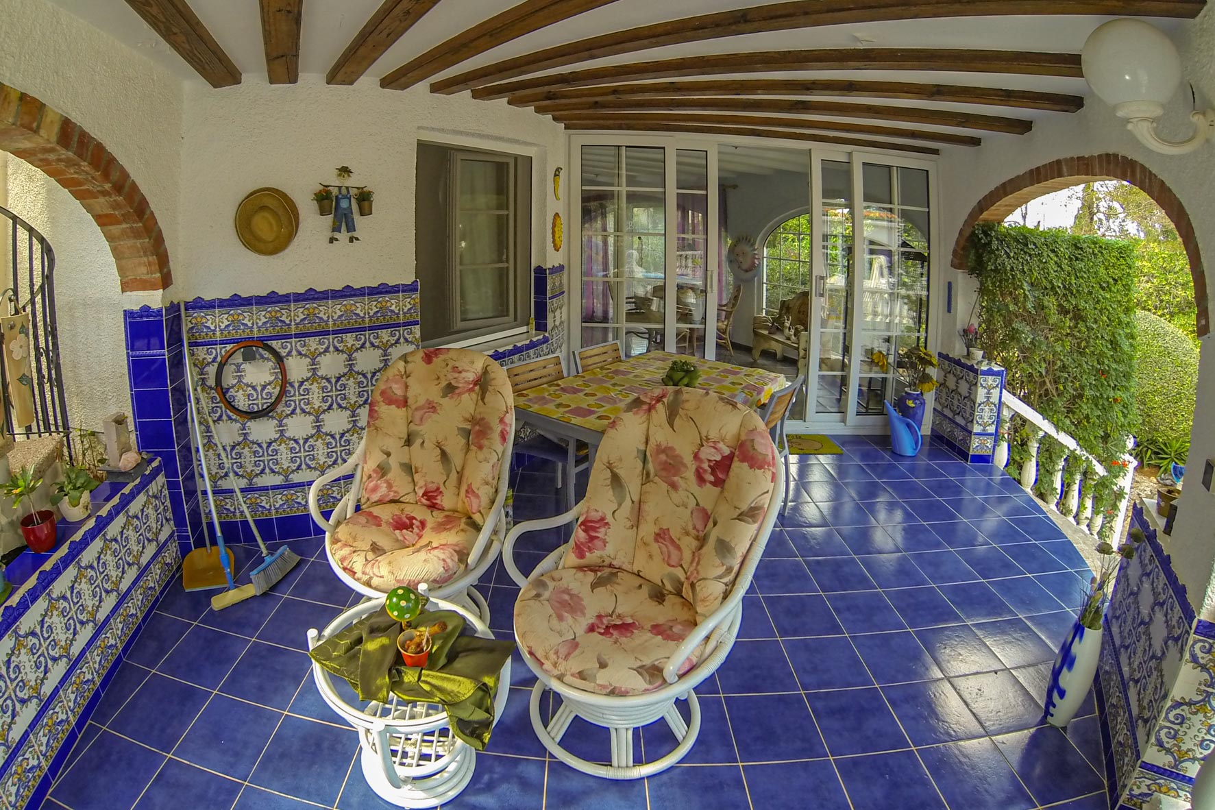 Overdekte veranda met blauwe tegels, bakstenen bogen en bloemrijke stoelen.