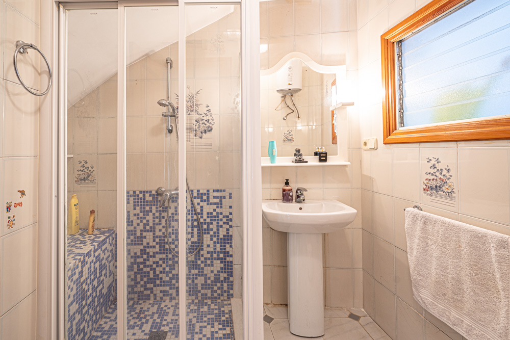 Badkamer met een inloopdouche afgewerkt met blauw mozaïek, een kolomwastafel en betegelde wanden met florale details en een houten raamkozijn.