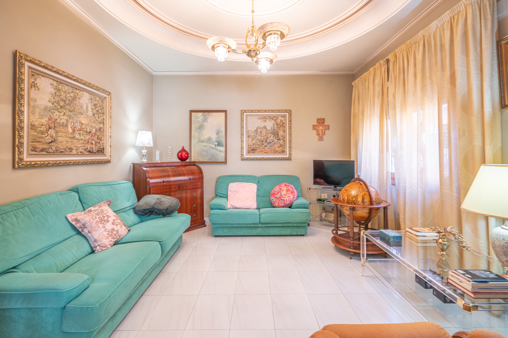 Lichte woonkamer met turquoise banken, witte tegelvloer en cirkelvormige plafondlijsten. Voorzien van klassieke houten meubels, een secretaire en ingelijste wanddecoraties.