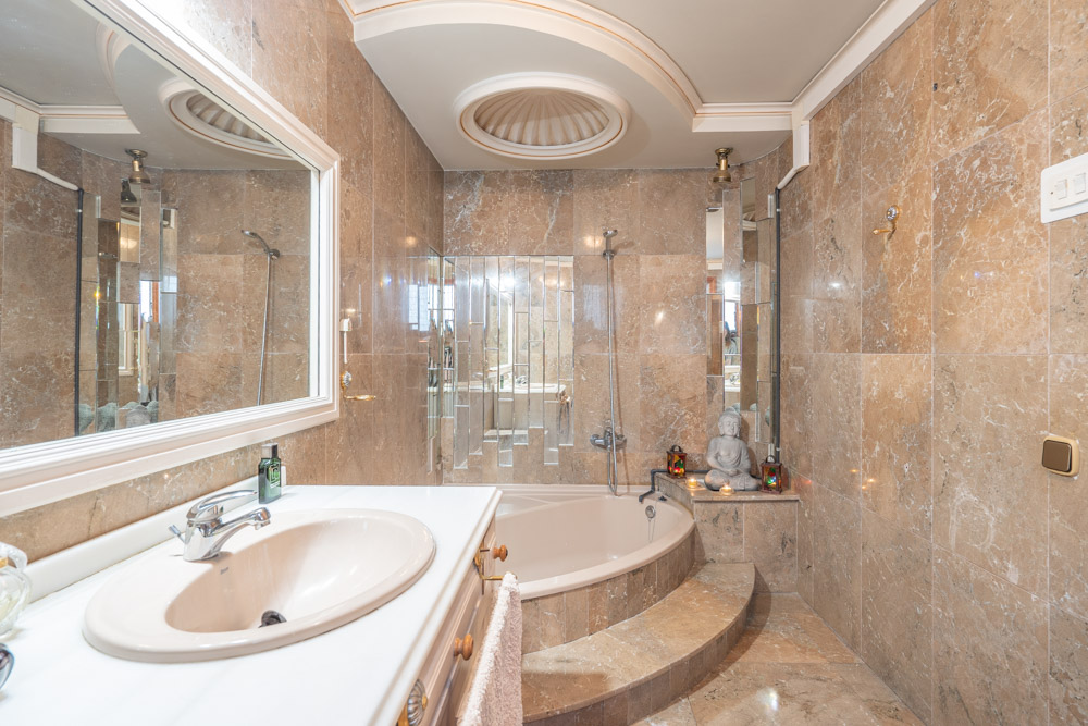 Marmeren badkamer met een ingebouwd ovaal ligbad, wit wastafelmeubel en een decoratief cirkelvormig plafonddetail.
