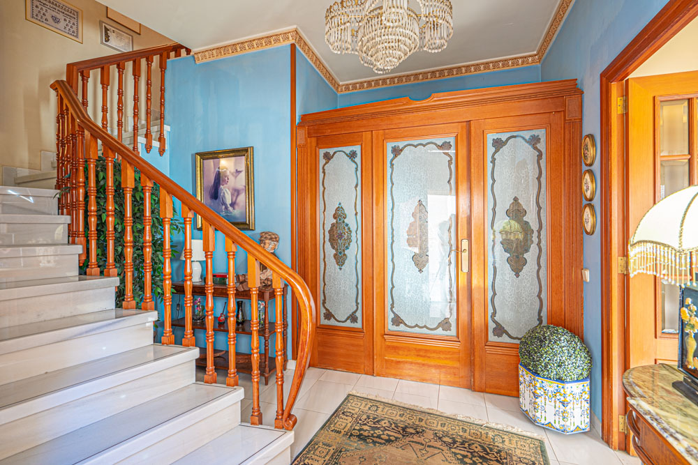 Ruime hal met een marmeren trap en houten balustrade. De ruimte bevat houten deuren met geëtst glas, sierlijsten aan het plafond en een klassieke kristallen kroonluchter.