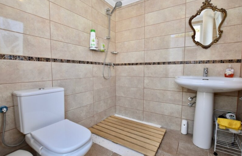 Badkamer met inloopdouche, houten vlonder mat, beige tegels met decoratieve rand en een klassieke spiegel met gouden lijst.