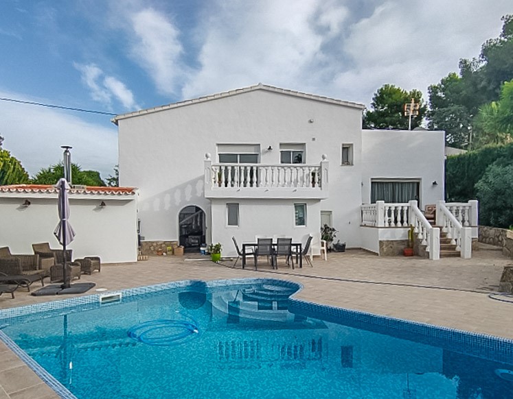 Buitenaanzicht van het blauwe mozaïekzwembad met een ruim betegeld terras, de witte gevel van de villa en diverse zitgedeelten in de open lucht.